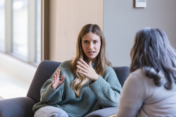A woman in a blue sweather sitting on a grey couch and explaning something to another woman with hand gestures