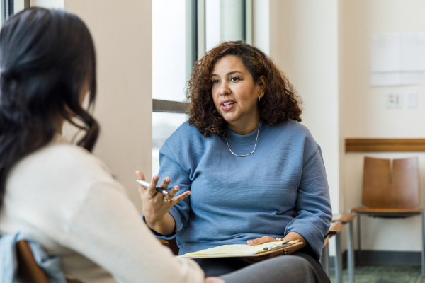 A woman in a blue top explaining something to another woman while holding a clipboard and gesticulating