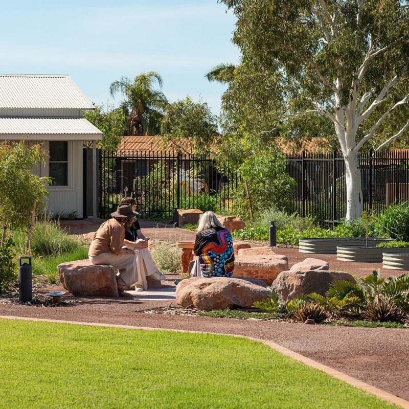 3 people sitting outside on rocks in a yarning circle surrounded by a garden